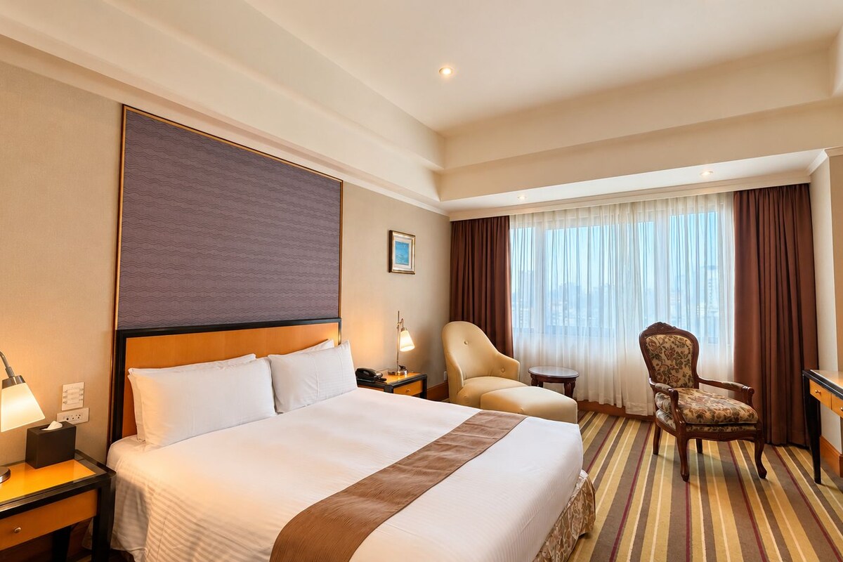 A hotel room with a large bed, a desk, two chairs, a nightstand with a lamp, and a window with curtains.