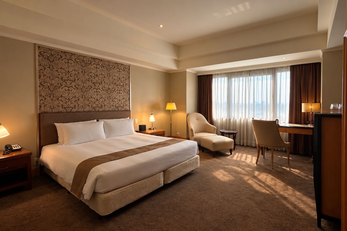 A hotel room with a large bed, two chairs, a desk, and a view of the city through the window.