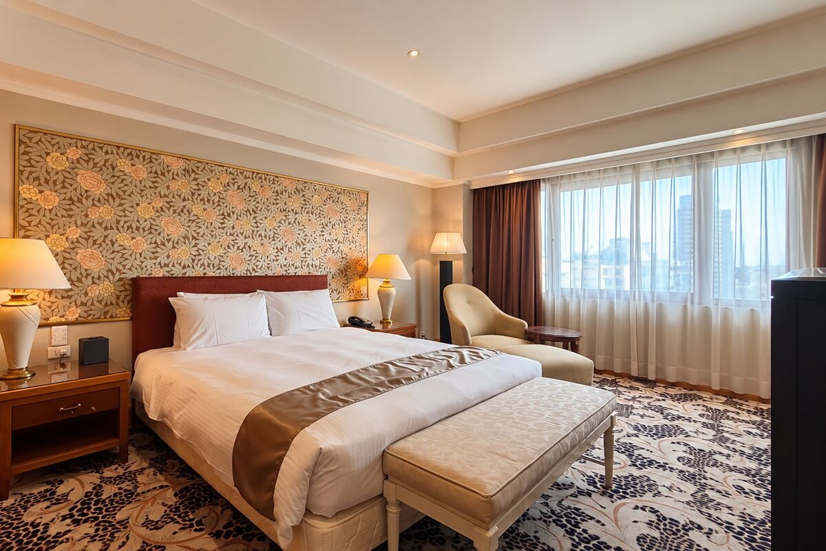 A hotel room with a large bed, bedside lamps, a chair, a desk, and a view of the city through the window.