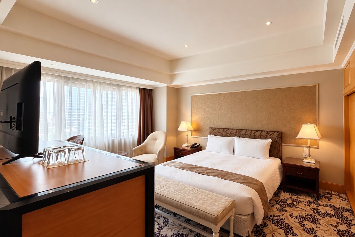 A hotel room with a bed, a desk with a television, a chair, two bedside tables with lamps, and a patterned carpet.