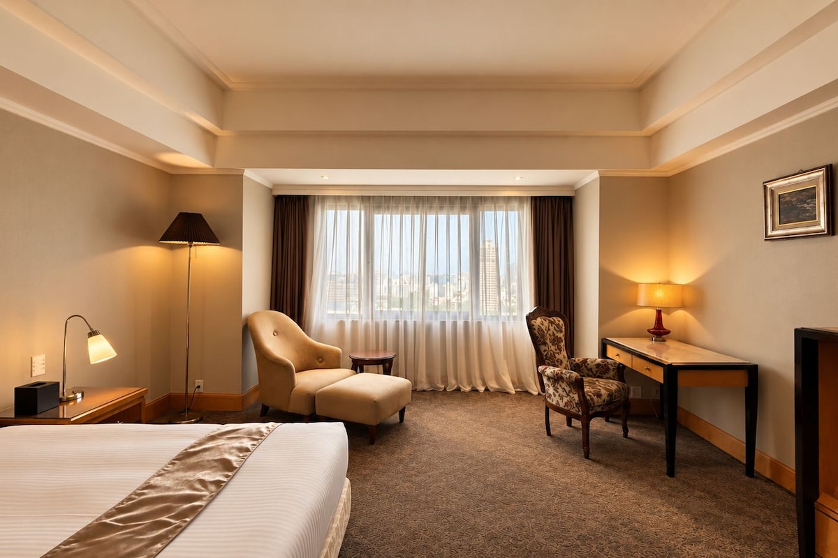 A hotel room with a bed, two armchairs, a desk, a lamp, and a view of the city through large windows.