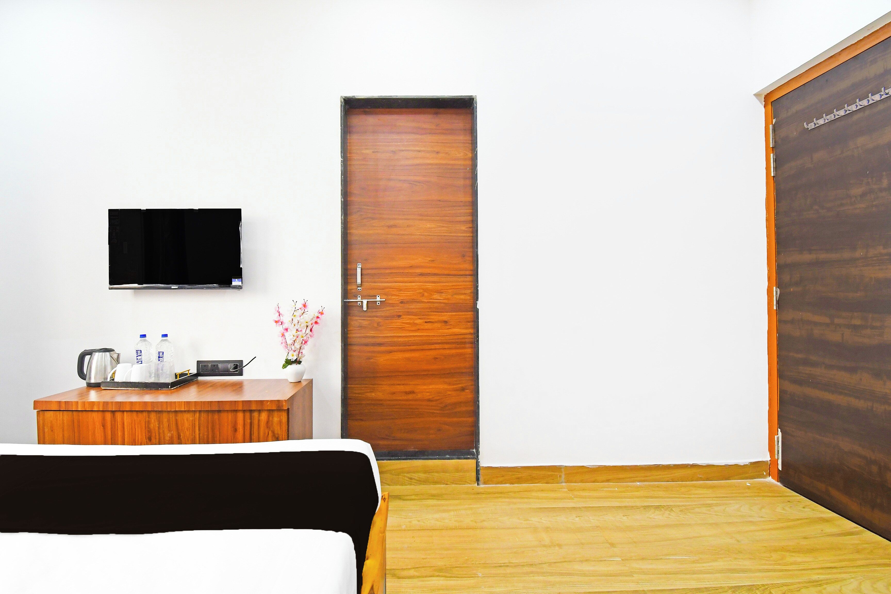 A hotel room with a bed, a wooden desk with a kettle, a flat-screen TV, and a wooden door.