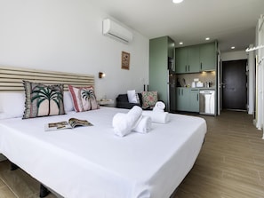 A hotel room with a large bed, a bedside table, a nightstand, a wall-mounted air conditioner, a kitchen area with cabinets and appliances, and a door leading to another room. - Sunstay Loft Saltillo (Torremolinos)