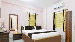 A bedroom with a large bed, a bedside table with a lamp, a dresser, a mirror, and an air conditioning unit. - Hotel O Ecopark Kolkata (Barasat)