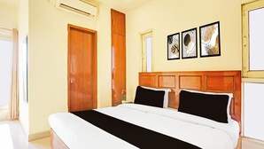 Hotellihuone, jossa on suuri sänky, puinen pääty, seinään asennettu ilmastointilaite, ovi ja kolme kehystettyä kuvaa.