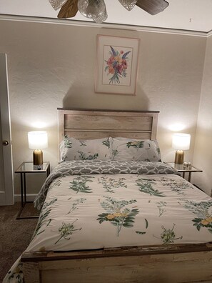 Un dormitorio con cabecera de madera, ropa de cama floral, dos lámparas de noche y un cuadro con motivos florales en la pared.