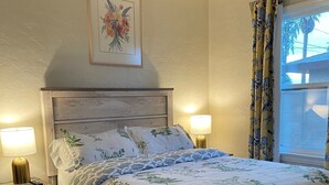 Een slaapkamer met een houten bed, twee bedlampen, een bloemistisch schilderij aan de muur en een raam met gordijnen.