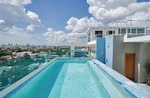 Une piscine à débordement située sur le toit, offrant une vue sur la ville, avec une balustrade en verre et un bâtiment moderne doté d’un mur en mosaïque bleue.