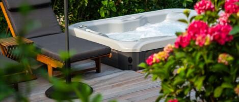 A hot tub on a wooden deck with lounge chairs and lush greenery in the background.