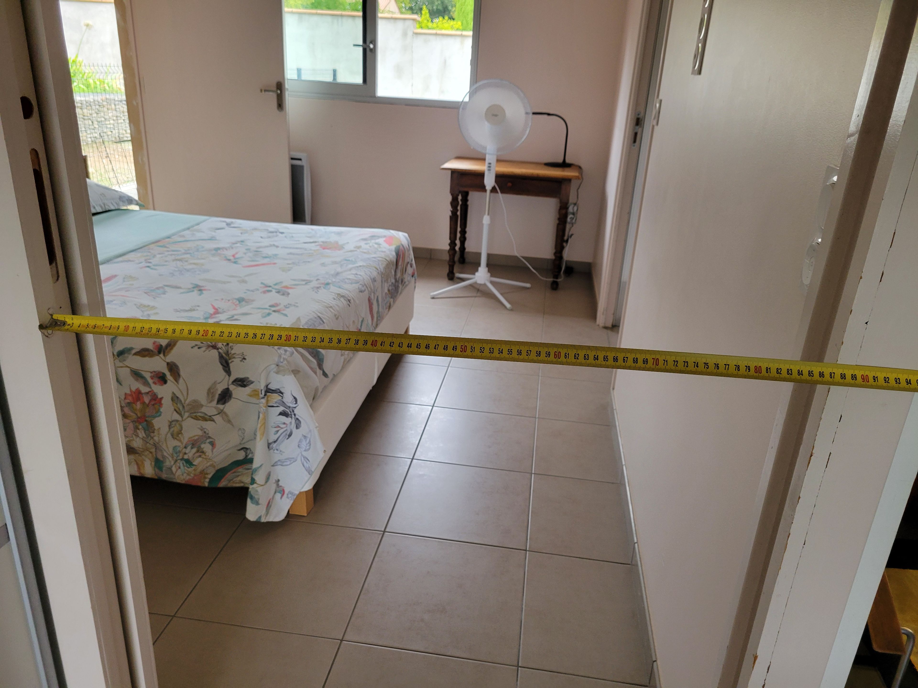 A bedroom with a bed, a standing fan, and a measuring tape across the door.