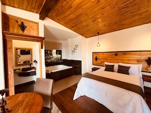 Ein Hotelzimmer mit Holzboden, einem großen Bett mit weißer Bettwäsche, einem hölzernen Kopfteil, einem kleinen Nachttisch, einem Spiegel und Blick auf ein Badezimmer mit einer Badewanne und Pflanzen.