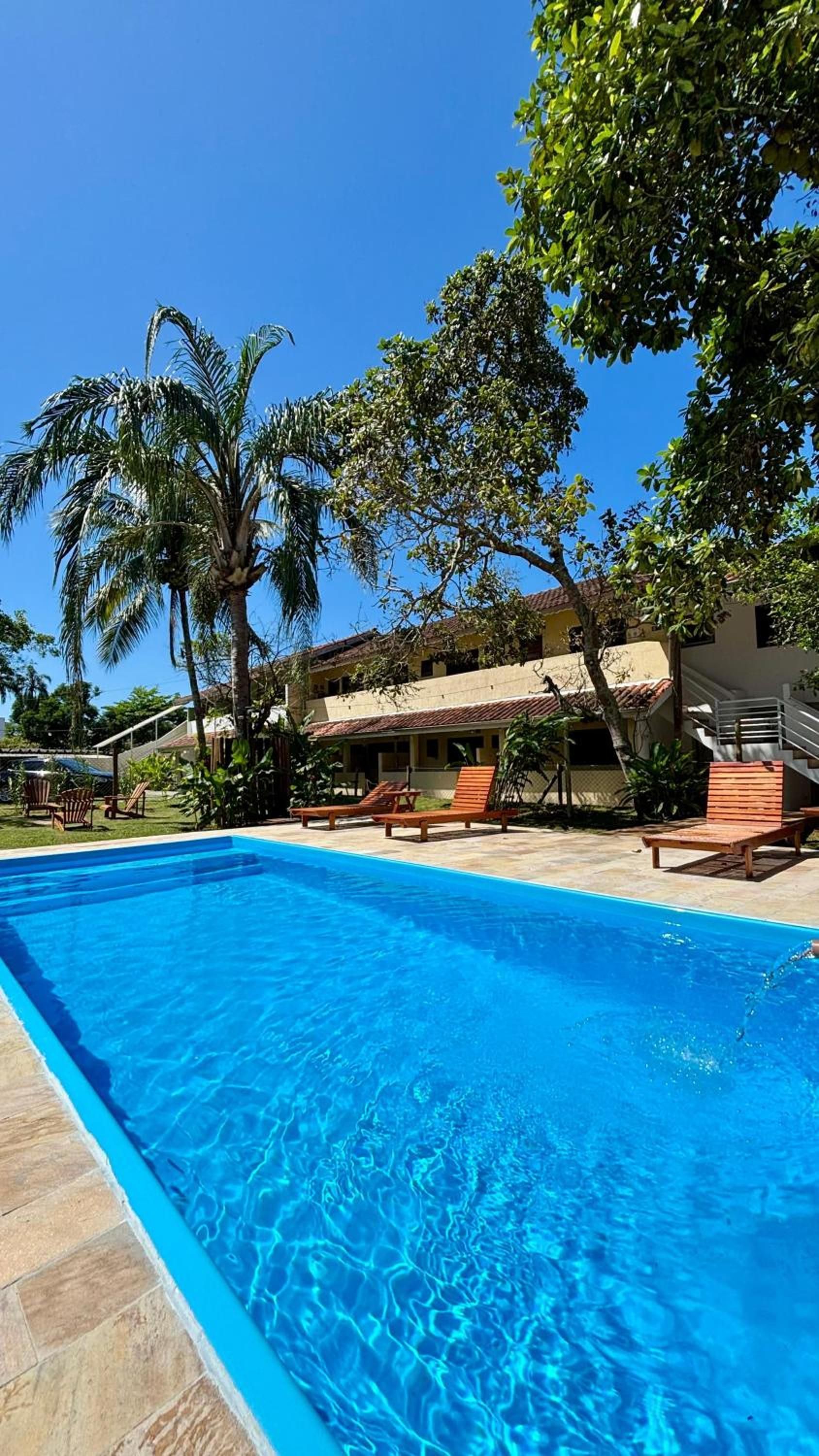 Piscina com água azul cristalina, cercada por espreguiçadeiras e palmeiras, com um edifício ao fundo.