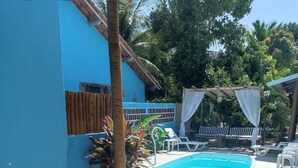 Une table et un banc en bois, placés dans un espace extérieur ensoleillé avec une piscine, le tout entouré de verdure et d’un bâtiment bleu.