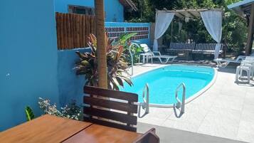 Mesa e banco de madeira em área externa ensolarada, com piscina, rodeados por vegetação e um edifício azul.