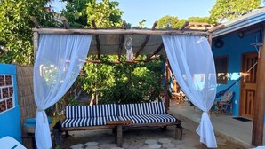 Un espace extérieur aménagé, abrité et doté de bancs et de fauteuils, entouré d’un bâtiment bleu et de verdure.