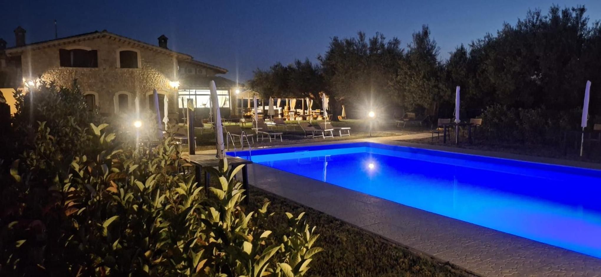 A swimming pool with lounge chairs and umbrellas, surrounded by greenery and a building with lights.