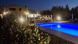 A swimming pool with lounge chairs and umbrellas, surrounded by greenery and a building with lights. - Villa Klinai (Cerveteri)