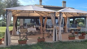 A covered outdoor seating area with tables and chairs, surrounded by potted plants. - Villa Klinai (Cerveteri)