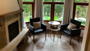 Een kamer met een open haard, twee zwarte stoelen, een ronde tafel en grote ramen met gordijnen.