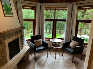 Una habitación con chimenea, dos sillas negras, una mesa redonda y ventanas grandes con cortinas.