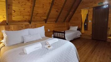 Uma cabana de madeira com duas camas, cada uma com roupa de cama e almofadas brancas, teto de madeira e uma porta com a inscrição 'SAIDA'.