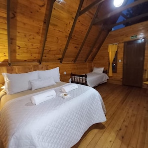 Uma cabana de madeira com duas camas, cada uma com roupa de cama e almofadas brancas, teto de madeira e uma porta com a inscrição 'SAIDA'.