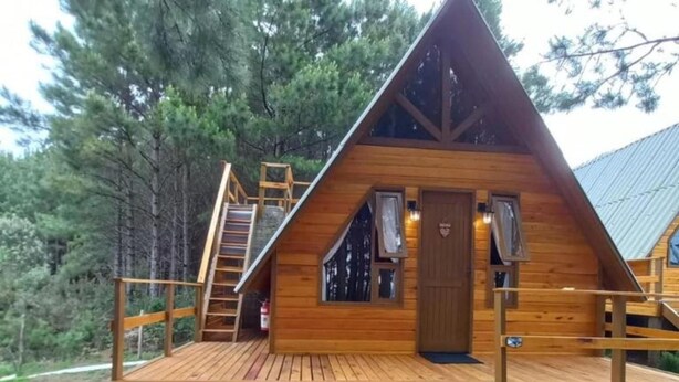 A wooden cabin with a steeply pitched roof, a wooden deck, and a staircase leading to the entrance. - Estalagem Refúgio Do Capão (Cambara do Sul)