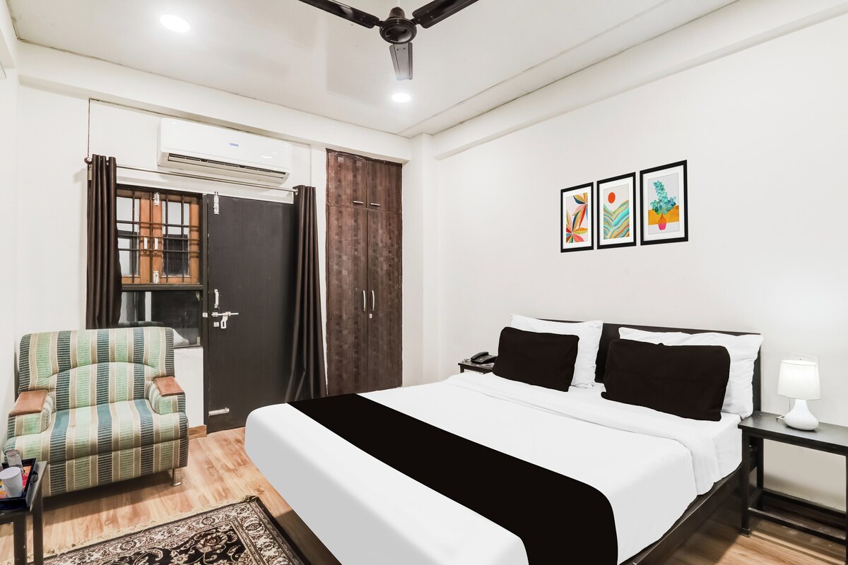 Quarto de hotel com cama, poltrona, guarda-roupa, mesa de cabeceira com abajur e parede com três obras de arte emolduradas.