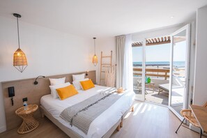 Une chambre avec un lit, des tables de chevet, une vue sur la plage et un balcon avec des sièges d’extérieur.