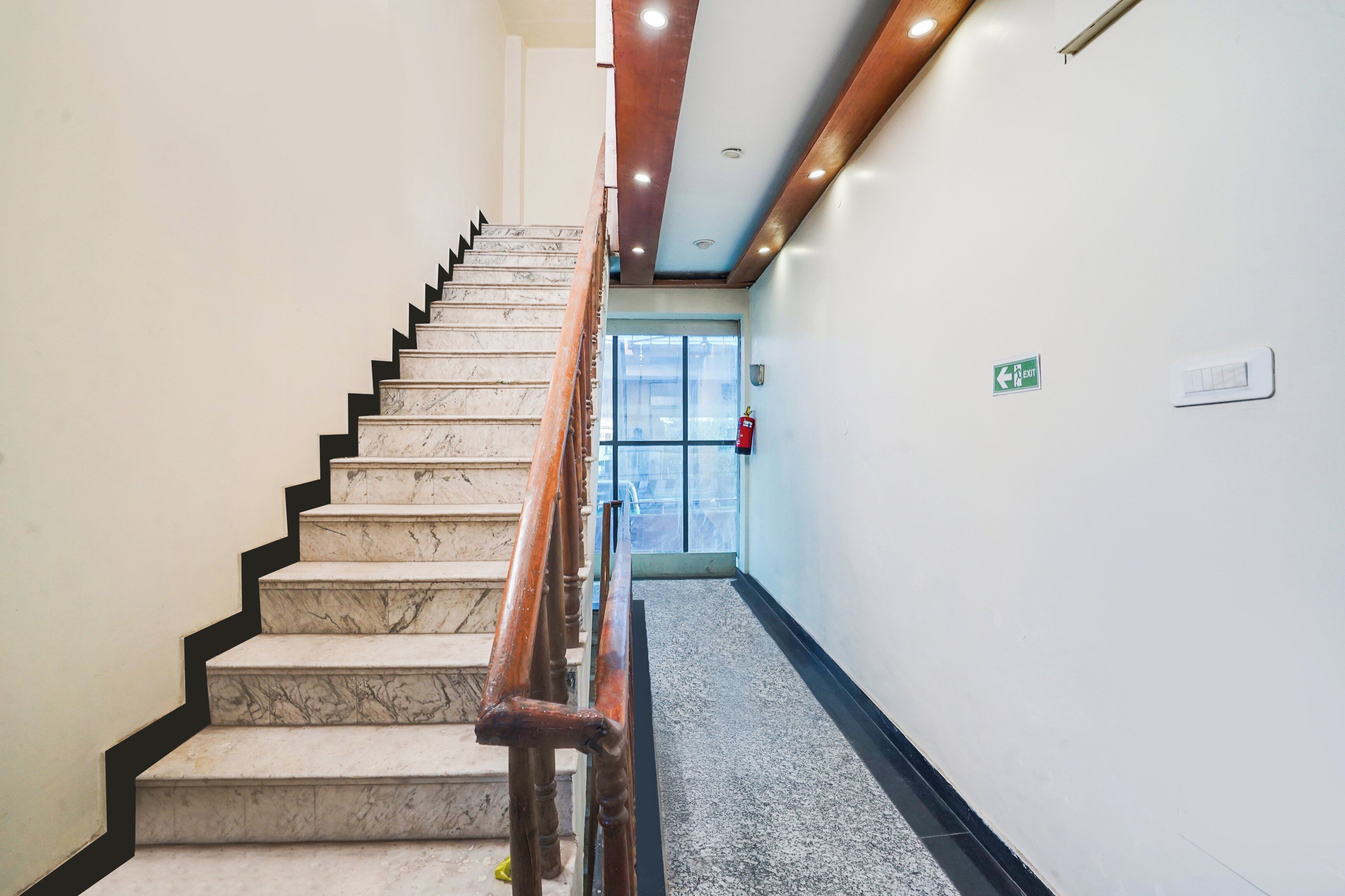 Escadaria de mármore com guarda-corpos de madeira que leva a uma porta com sinalização de saída.