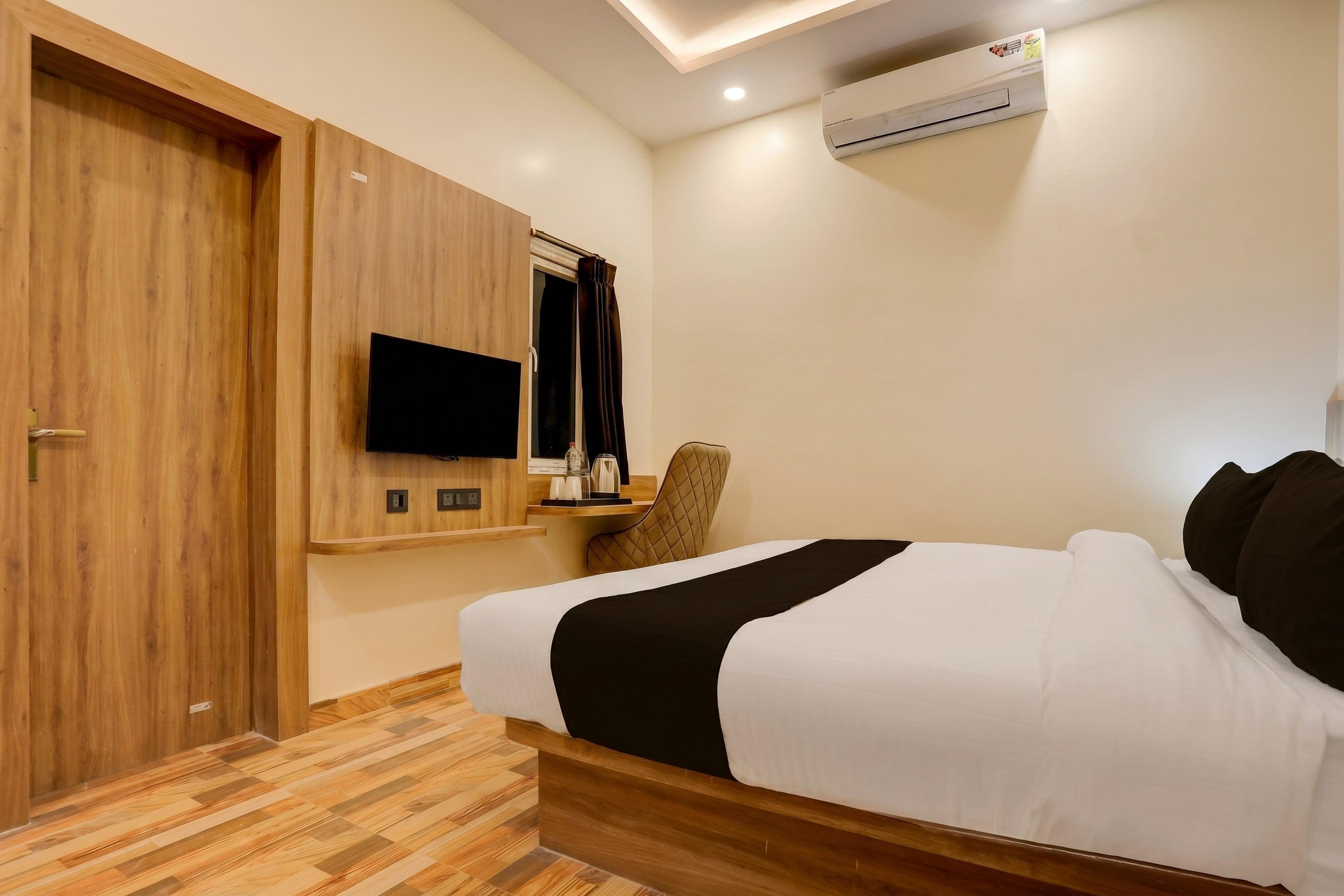 A hotel room with a bed, a desk with a chair, a flat-screen TV, and an air conditioning unit.