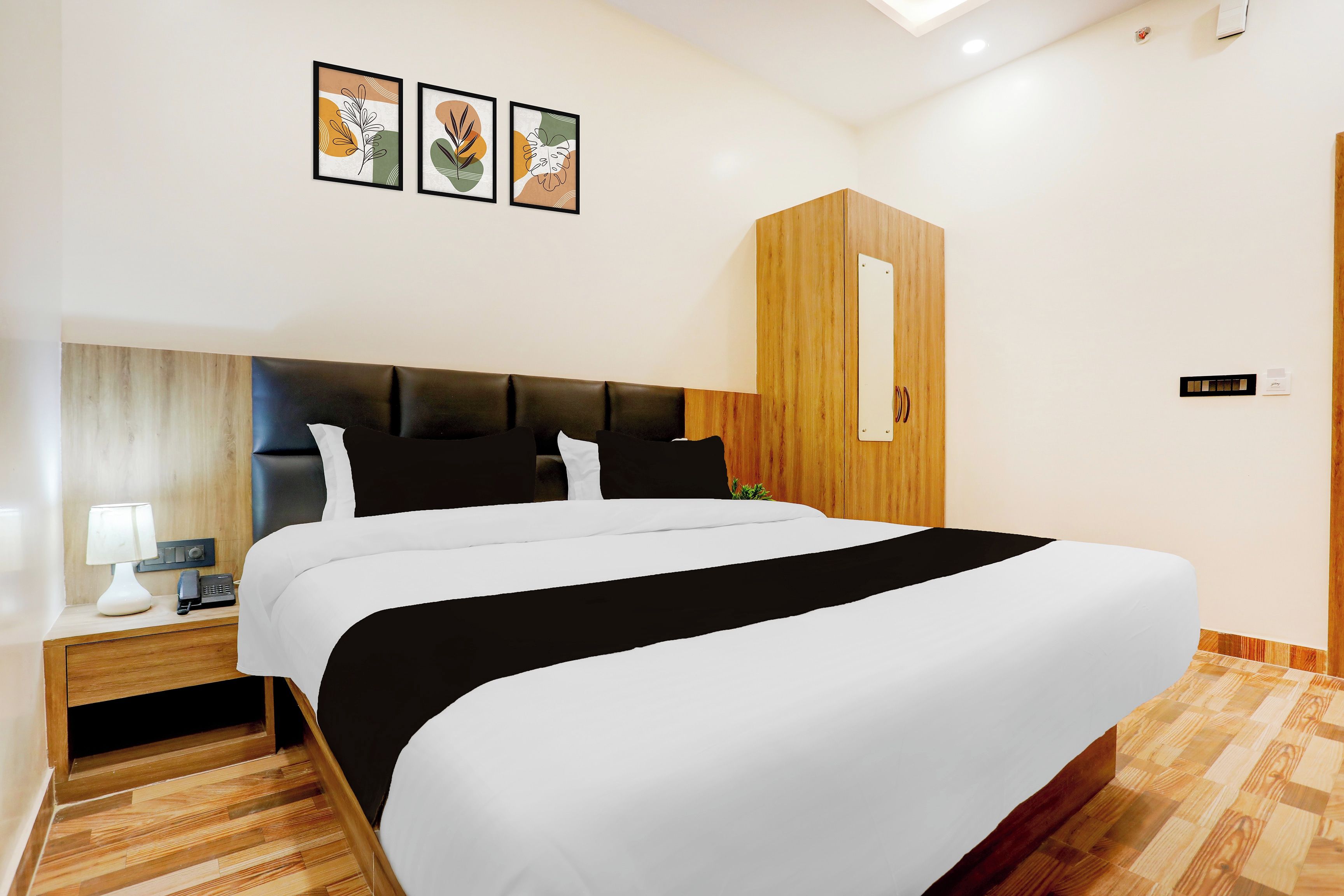 Una habitación de hotel moderna con una cama grande, cabecera de madera, mesita de noche con lámpara y una planta pequeña.