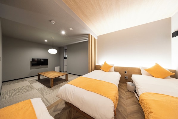 A hotel room with two beds, a small table, a flat-screen TV, and a bedside table with a lamp.
