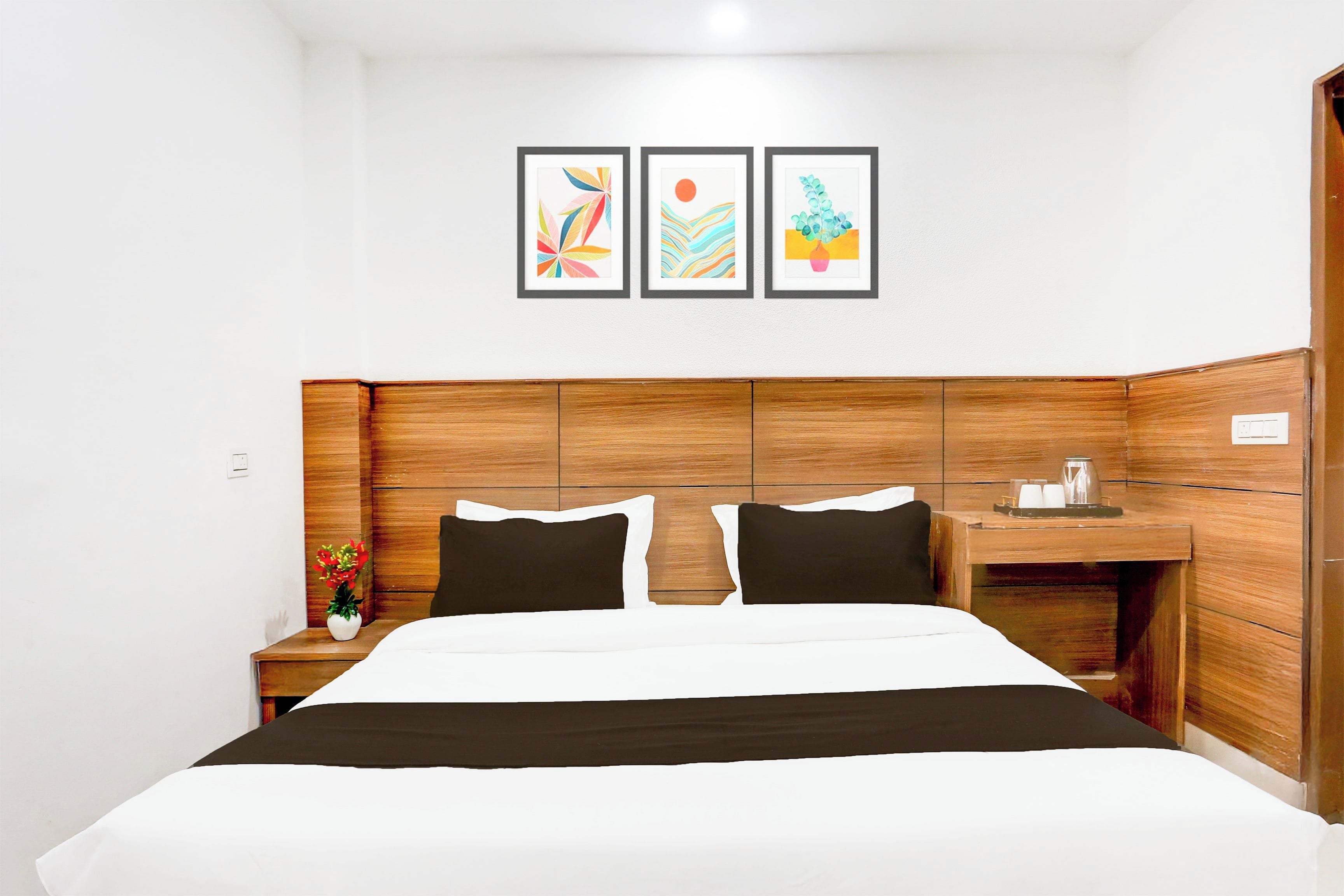 A hotel room with a wooden headboard, a bed with black and white bedding, a nightstand with a vase of flowers, and three framed artworks on the wall.