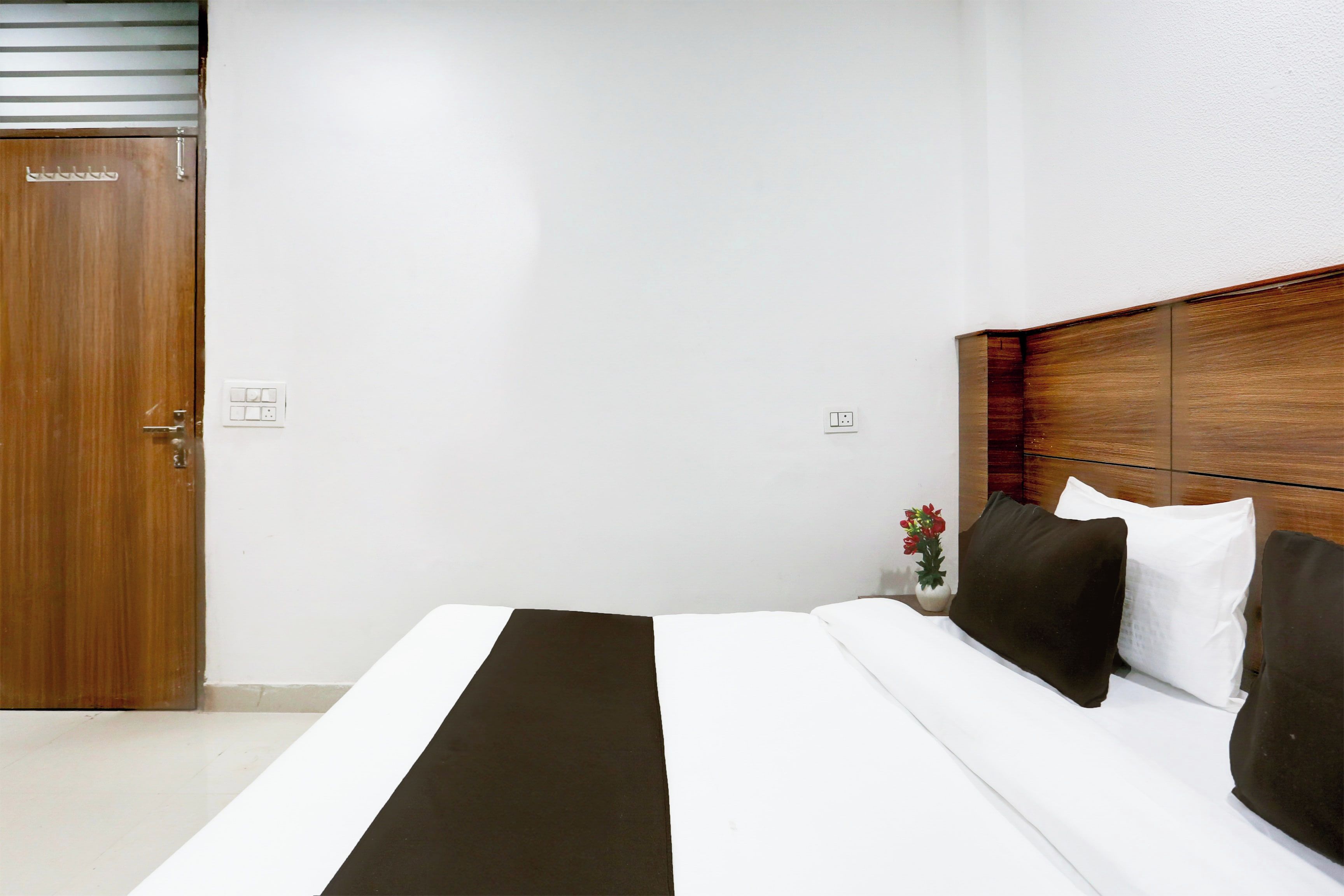 A hotel room with a bed, a wooden headboard, a nightstand with a vase of flowers, and a door with a coat rack.