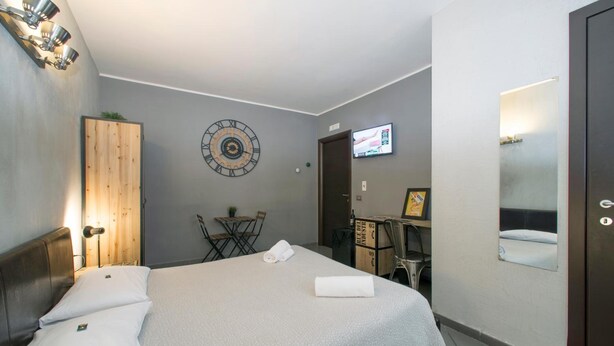 A hotel room with a bed, a wooden wardrobe, a wall clock, a television, and a desk with a chair. - Il Taschino Suite (Naples)