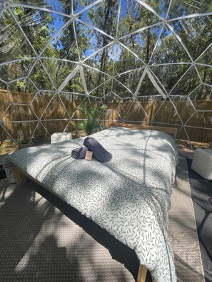 Una cama con una colcha estampada, un almohadón y una toalla enrollada en una habitación con una estructura de techo geométrica y paredes de madera.