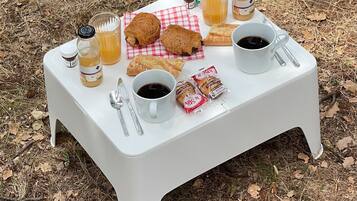 Un coin pique-nique aménagé avec une table blanche, deux tasses de café, un verre de jus de fruits, des croissants et des en-cas emballés, le tout disposé sur une nappe à carreaux à l'extérieur.