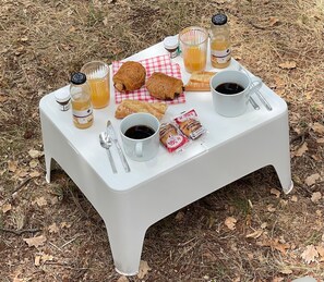 Un montaje para picnic con una mesa blanca, dos tazas de café, un vaso de jugo, croissants y aperitivos envasados sobre un mantel a cuadros al aire libre.