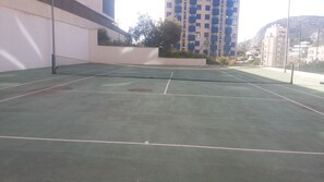 En udendørs tennisbane med net, omgivet af bygninger og et bjerg i baggrunden.