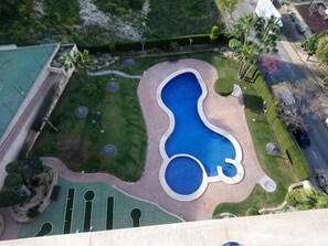 Ovenfra ses en swimmingpool med en usædvanlig form, omgivet af velplejet græs og træer.