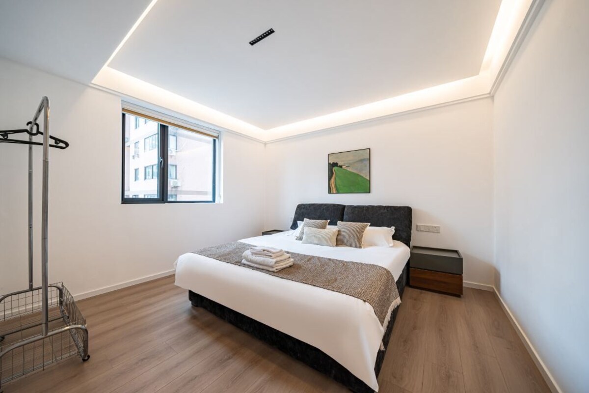 Ein modernes Schlafzimmer mit einem großen Bett, einem Fenster, einem Gemälde und einem Metallwagen.
