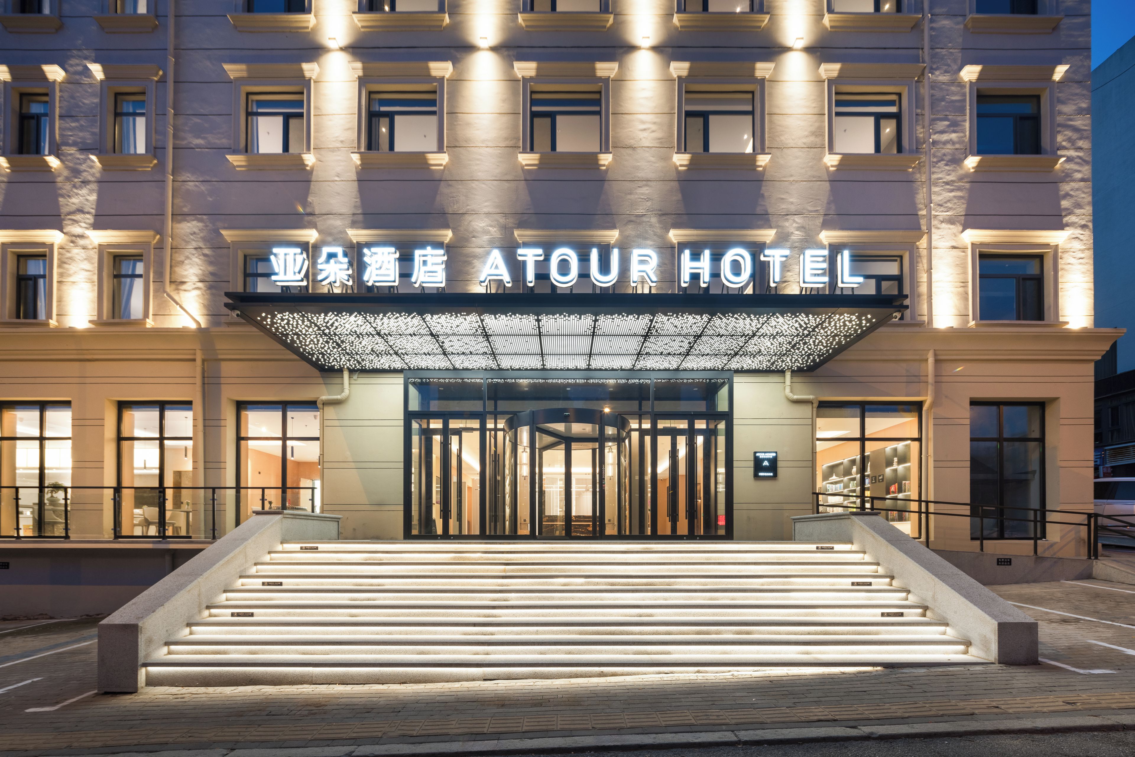 Entrada de hotel con una escalinata monumental, un gran letrero iluminado y una fachada moderna.