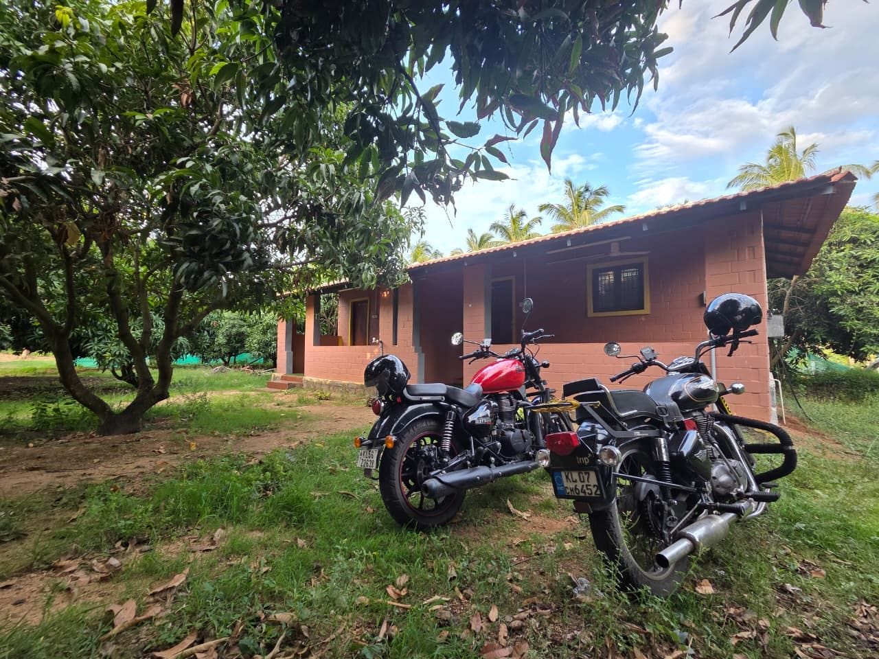Dos motocicletas estacionadas frente a un edificio de ladrillo con techo rojo, rodeado de follaje y árboles.