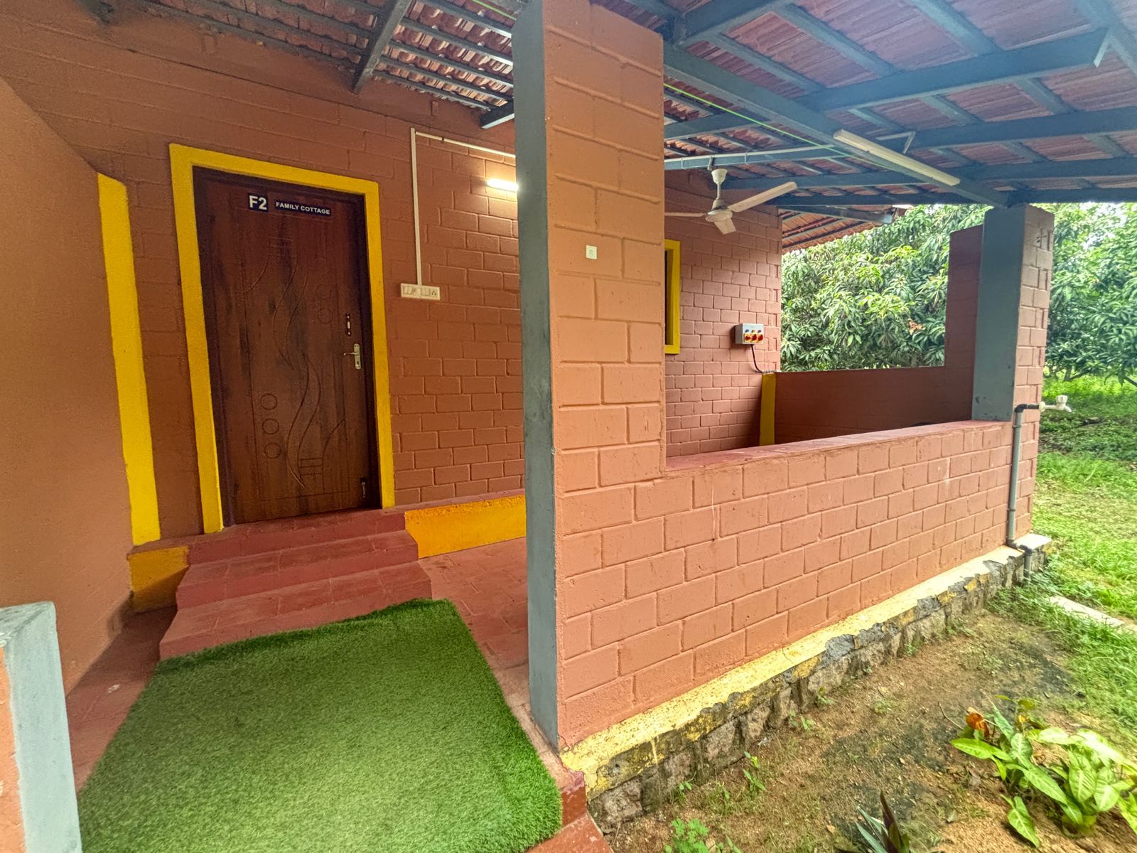 Un espacio exterior techado con pared de ladrillo, una puerta de madera con la etiqueta 'F2 FAMILY COTTAGE' y un pequeño parterre de césped.