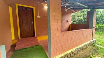 Un espacio exterior techado con pared de ladrillo, una puerta de madera con la etiqueta 'F2 FAMILY COTTAGE' y un pequeño parterre de césped.