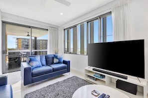 Een moderne woonkamer met een grote flatscreen televisie, een blauwe bank met gekleurde kussens, en een uitzicht over de stad door grote ramen.