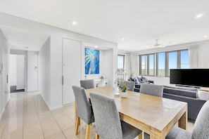 A modern living room with a dining area, a flat-screen TV, and a large window with a view. - Deja Blue – Your Serenity Short Stays Darwin Waterfront Escape (Darwin)