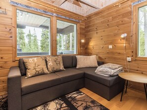 Interior d'una cabana de fusta amb un sofà, almoinades, una taula petita i una finestra amb vista a arbres.