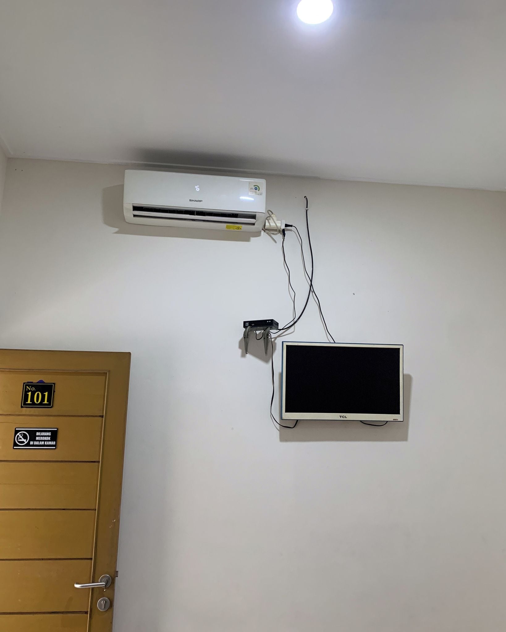 Una habitación con un aire acondicionado blanco, un televisor montado y una puerta con un cartel que indica el número de habitación 101.